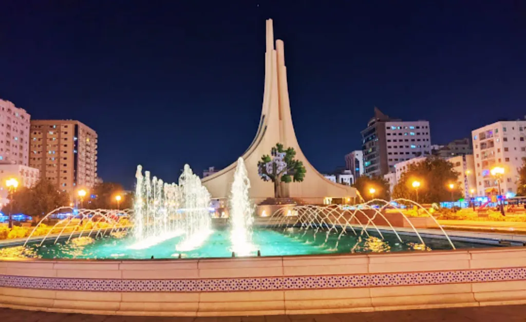Nighttime view of the iconic Martyrs' Memorial in Algiers, Algeria, with illuminated fountains in the foreground and city lights in the background.