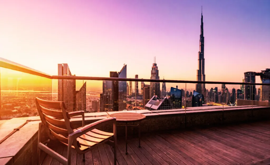 Rooftop terrace overlooking a modern city skyline at sunset. Wooden chair and table in foreground, with skyscrapers and vibrant purple-orange sky. Calm ambiance