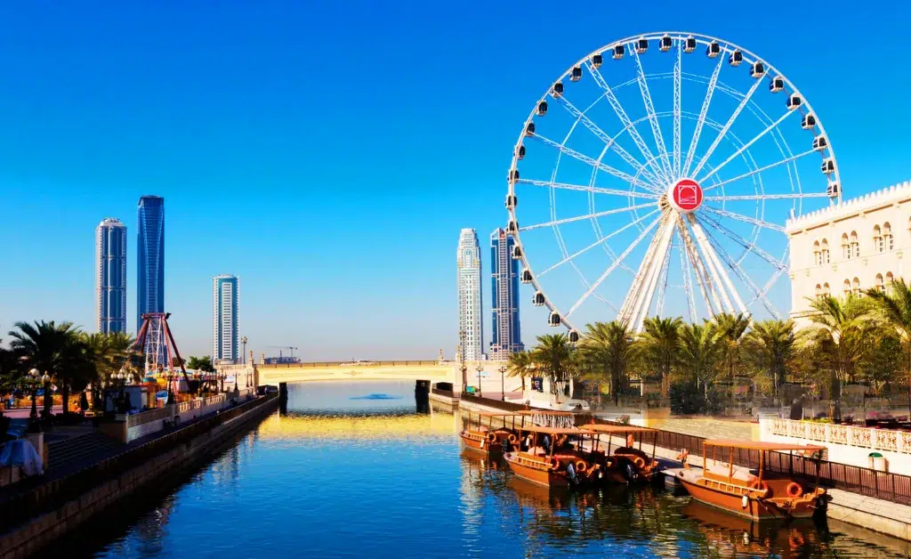 A vibrant cityscape featuring a large Ferris wheel next to a serene river lined with palm trees. Skyscrapers loom in the background under a clear blue sky.