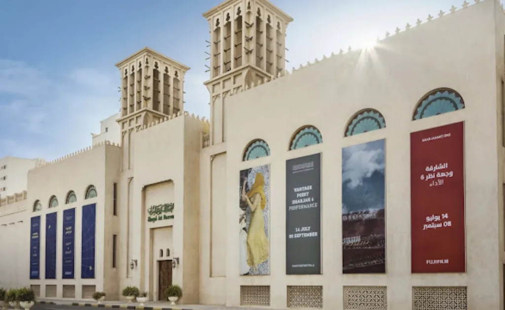 A beige building with traditional wind towers under a clear sky features large, colorful banners displaying art and event details, creating a vibrant atmosphere.