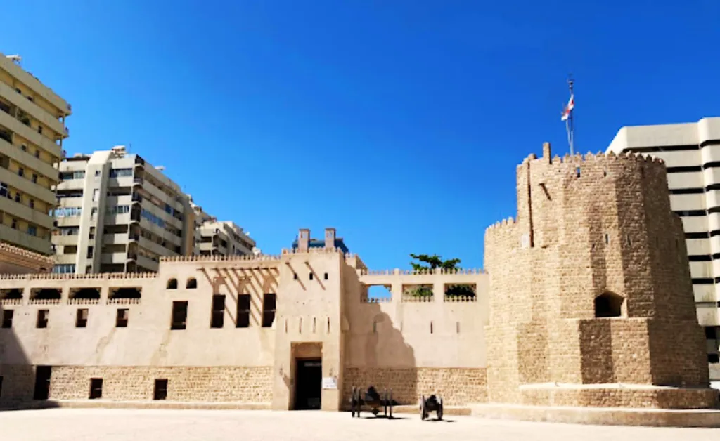 A historic fort with beige stone walls stands under a clear blue sky. Modern buildings flank it, creating a contrast between old and new architecture.