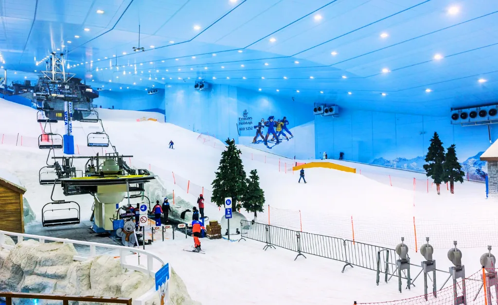 Indoor ski resort scene with a snowy slope, skiers, and a ski lift. Bright lights illuminate the space, giving a lively and inviting atmosphere.
