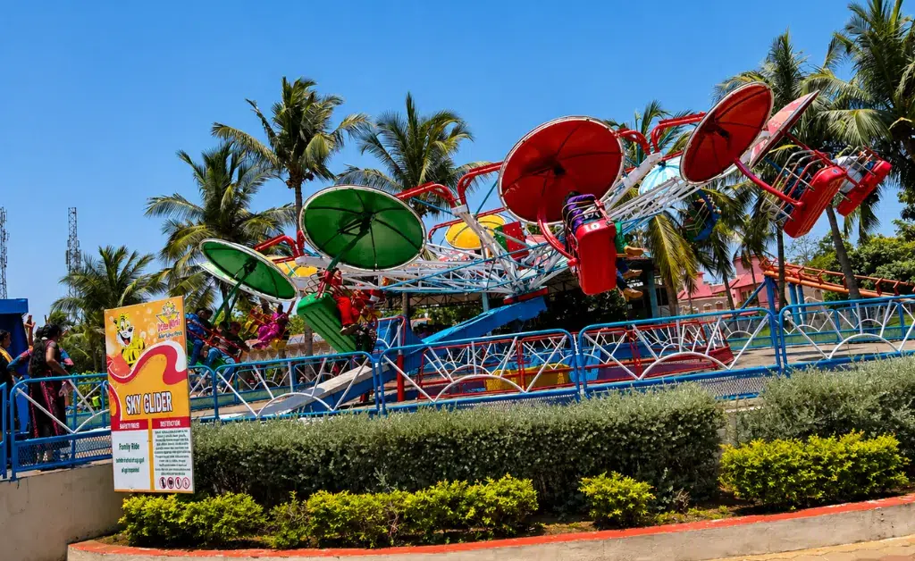 Colorful amusement park ride with circular seating swings tilting, surrounded by palm trees under a bright blue sky. A "Sky Glider" sign in the foreground.