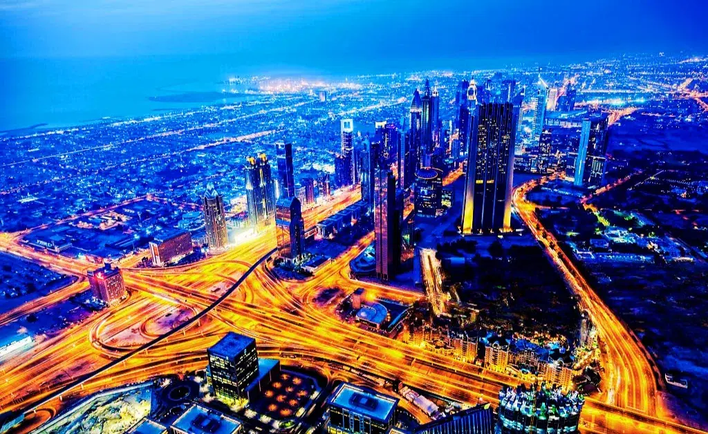 A vibrant aerial view of a cityscape at night, featuring illuminated skyscrapers and roads radiating bright, glowing lights, creating a lively urban ambiance