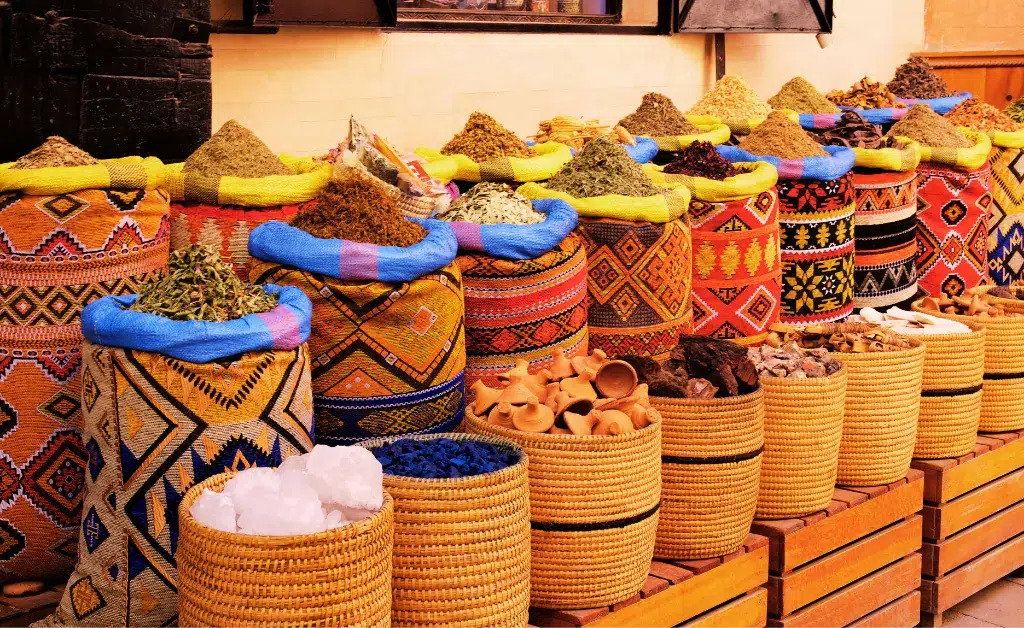 Vibrant market scene with woven baskets full of colorful spices and herbs, arranged neatly in rows. Decorative patterns enhance the lively atmosphere.
