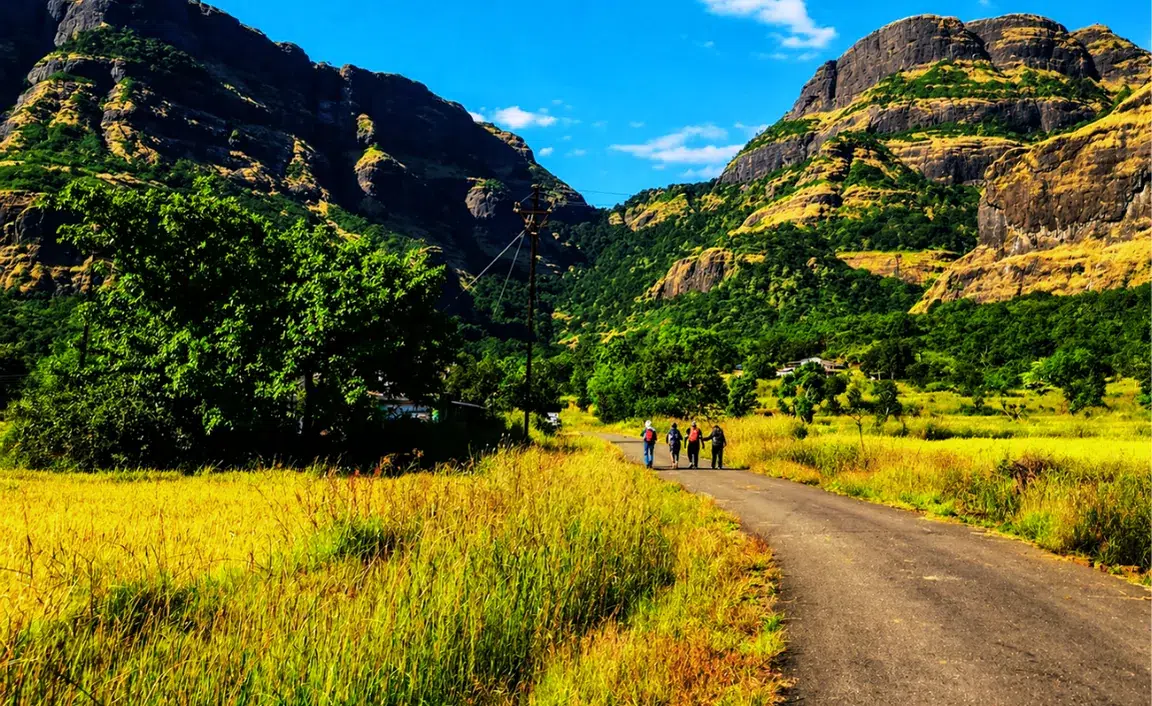 A group of people walks down a winding road through a lush, sunlit valley with towering, green mountains in the background under a clear blue sky.