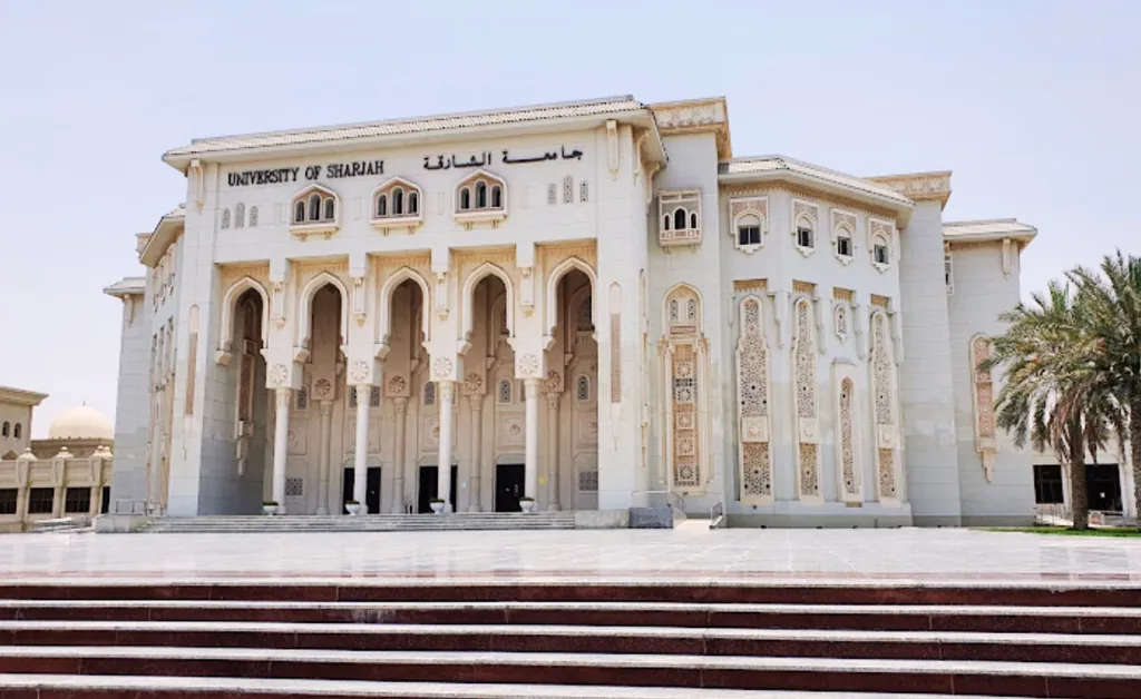 Grand facade of University of Sharjah with ornate arches and pillars against a clear sky, palm tree visible on the right, conveys grandeur.