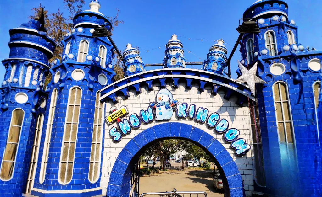 The image depicts the vibrant entrance of Snow Kingdom, featuring blue towers and a snowman sign above the archway. The overall tone is playful and inviting.