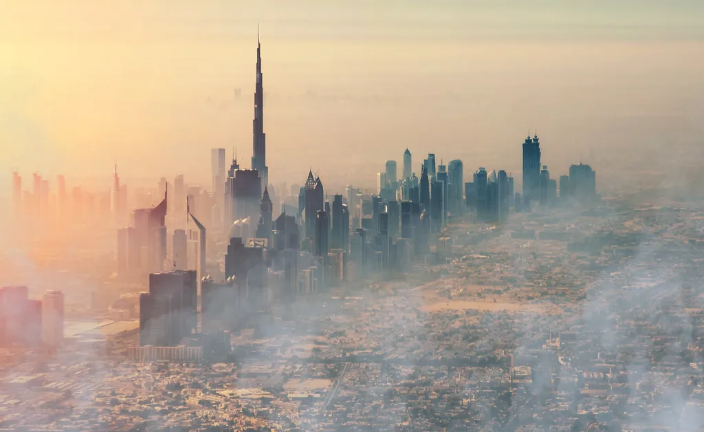 Aerial view of a hazy city skyline at sunset, with a tall skyscraper dominating the scene. Buildings cast long shadows, exuding a tranquil, urban atmosphere.