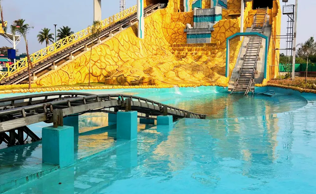 A vibrant water park features a yellow and orange water slide structure with cascading blue water. Tracks cross a clear turquoise pool, evoking a fun atmosphere.