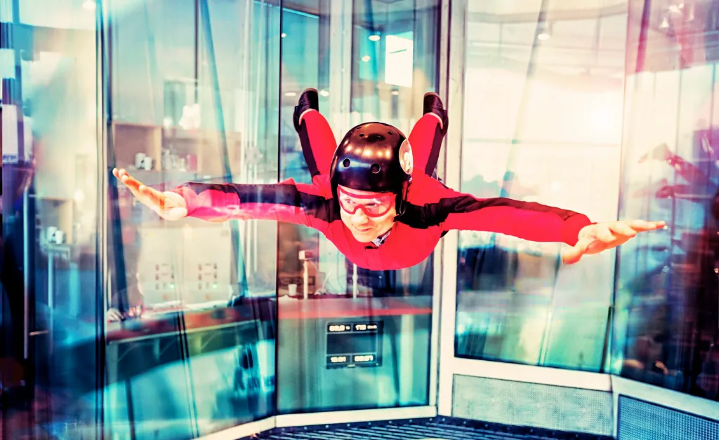 A person in a red and black suit and helmet is indoor skydiving, floating horizontally in a wind tunnel. The scene conveys excitement and thrill.