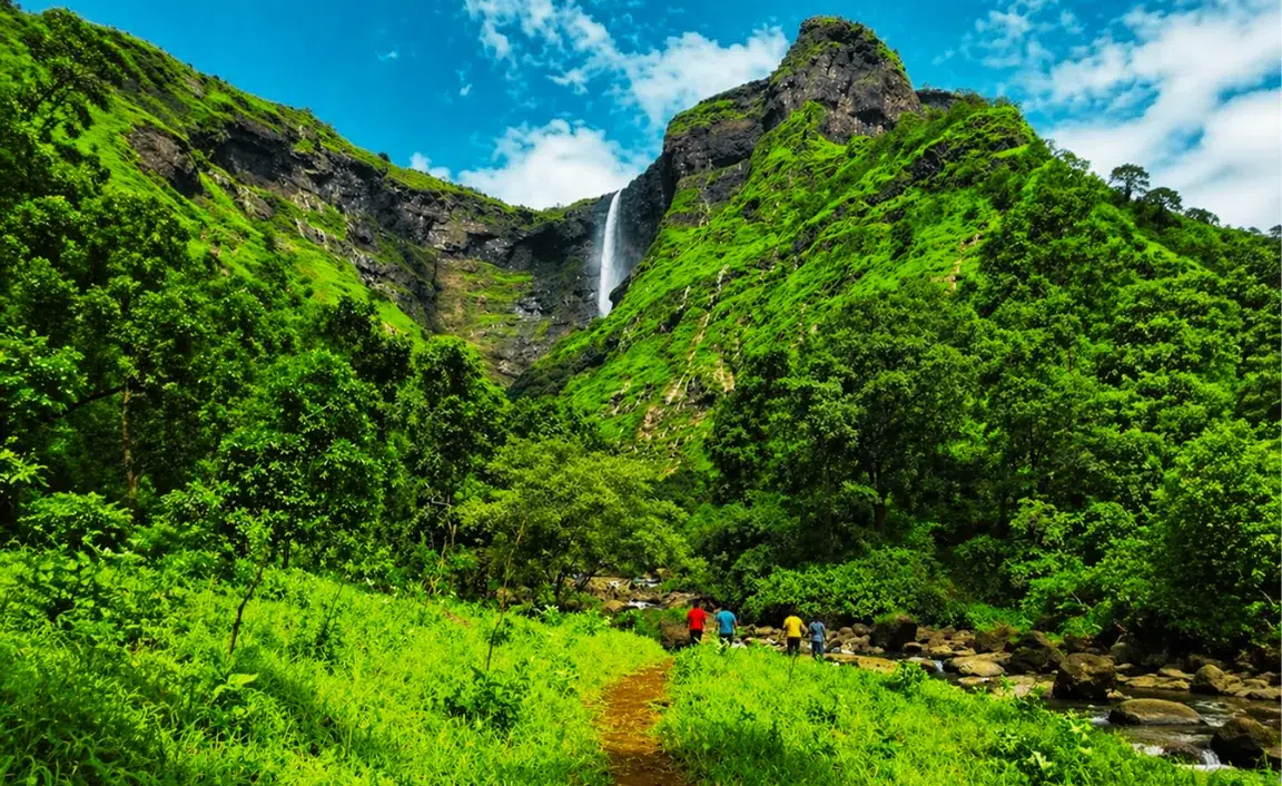 A lush green landscape with a tall waterfall cascading down a rocky mountain. Five hikers in colorful clothing walk along a dirt path toward the waterfall under a bright blue sky.