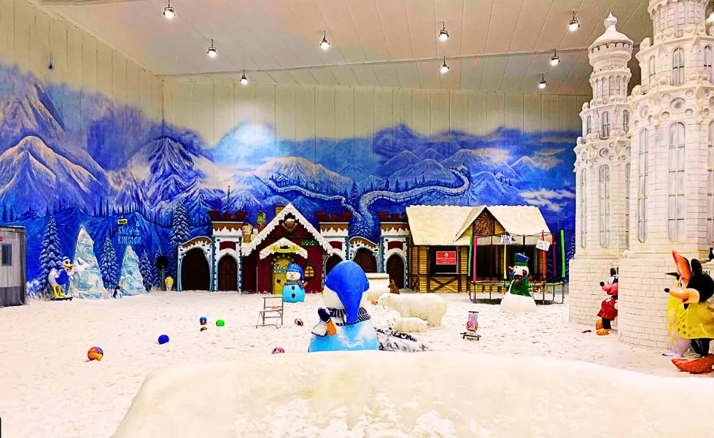 Winter-themed indoor park with artificial snow, colorful snowman, cartoon characters, castle, and village backdrop. Playful, festive atmosphere.