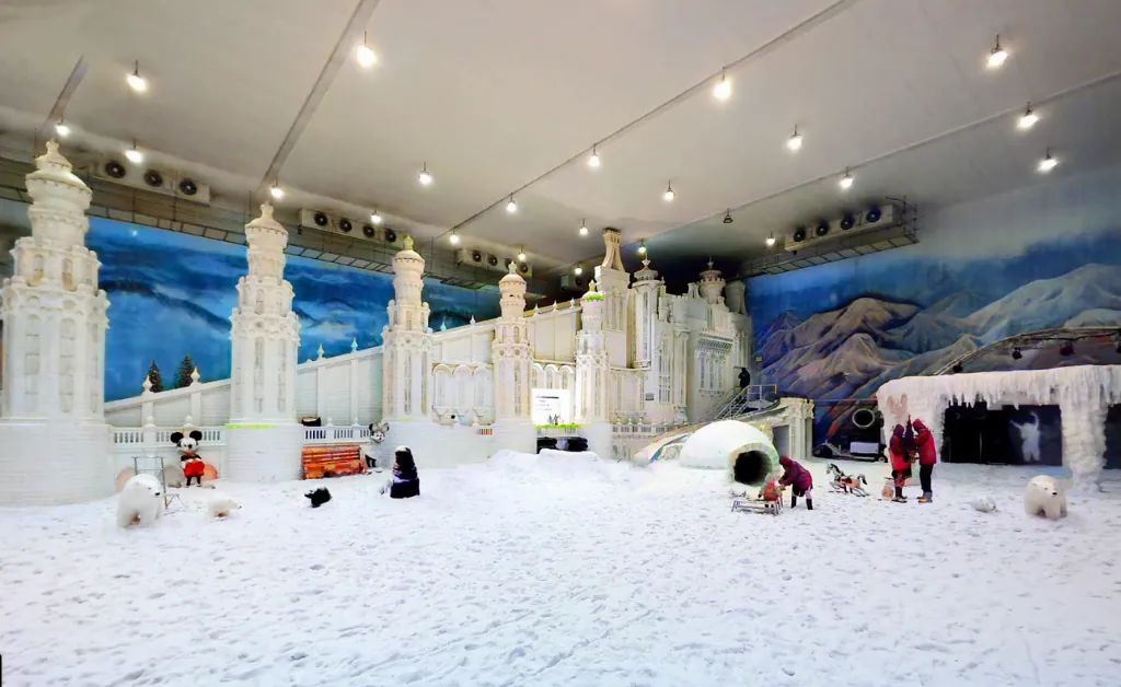 Indoor theme park with snowy décor, featuring a grand castle, igloo, and artificial snow. People enjoy activities, creating a festive, wintry atmosphere.
