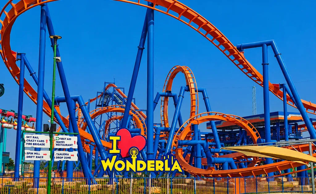A bright, clear landscape view of a colorful roller coaster at an amusement park, featuring orange tracks looping around blue support beams, with a large “I ❤️ Wonderla” sign in the foreground and no people present. Wonderla Chennai