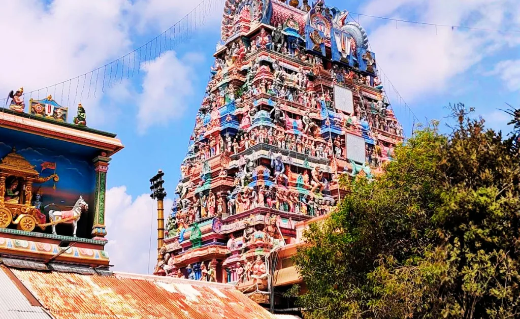 A vibrant Hindu temple tower with intricate carvings and colorful statues against a blue sky. Lush greenery surrounds the scene, evoking a serene atmosphere.
