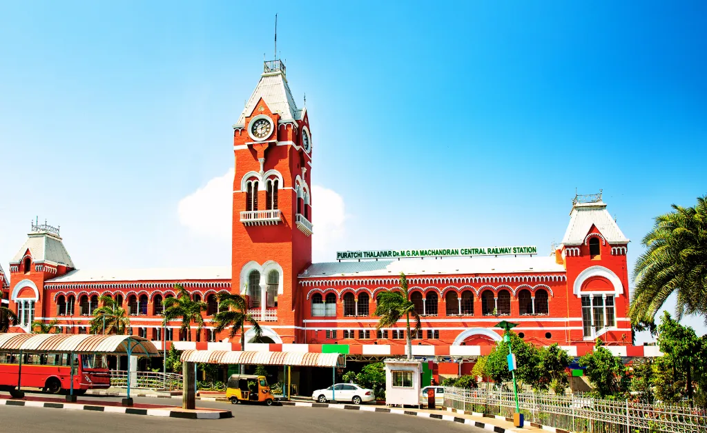 The image shows a vibrant red colonial-style railway station with a clock tower under a clear blue sky. Buses and trees are visible in the foreground.
