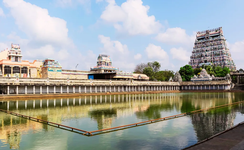 Grand temple with ornate towers reflecting in a large, serene water tank under a bright blue sky with fluffy clouds, conveying peace and grandeur.