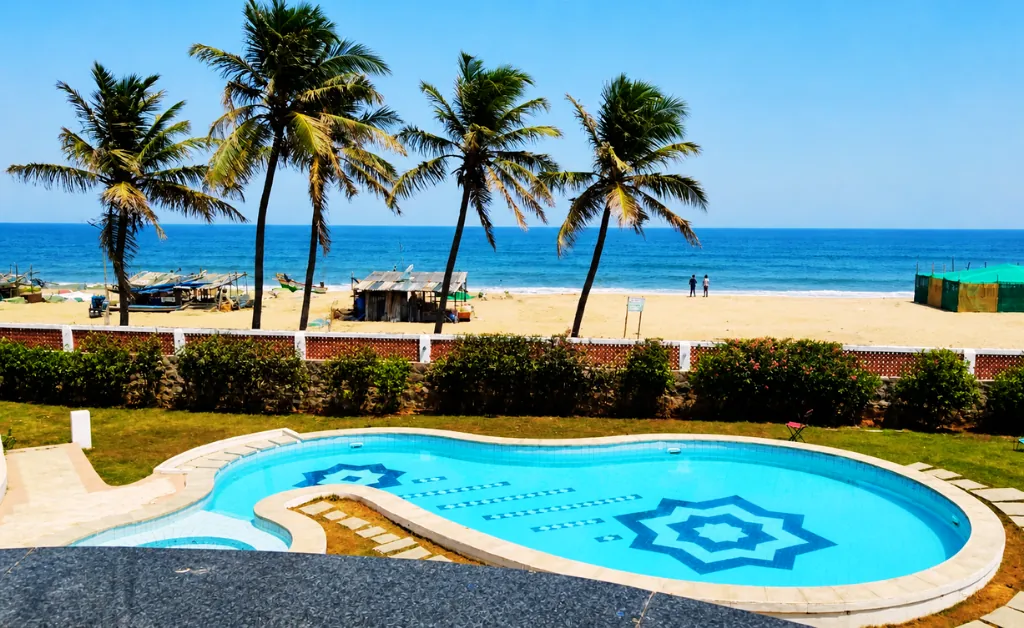Curved pool with geometric design overlooks a sandy beach with palm trees. Calm ocean and clear blue sky create a serene, tropical atmosphere.