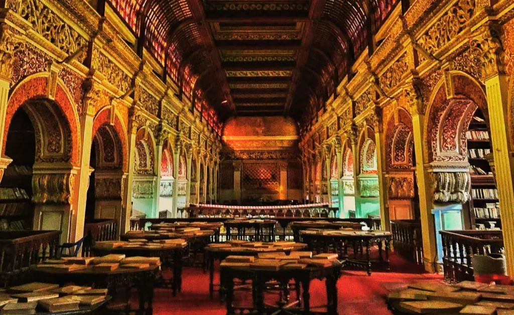 Ornate, historic library interior with arched ceilings, intricate carvings, wooden bookshelves, and tables with scattered books, evoking a majestic, intellectual ambiance.