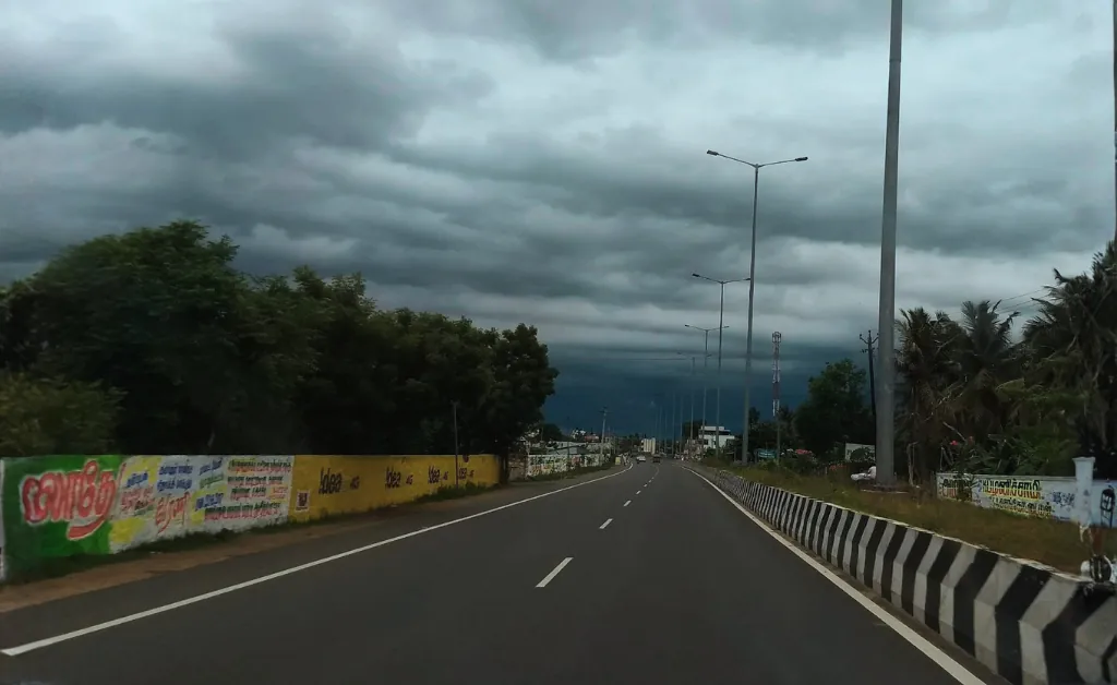 A deserted highway stretches ahead under a dark, cloudy sky, flanked by trees and colorful billboards, creating a moody, imminent rain atmosphere.