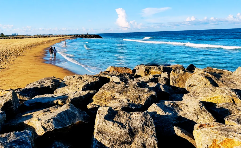 Rocky shore with large boulders, adjacent to a sandy beach. Two people walk along the shoreline of the calm, blue ocean under a bright sky.