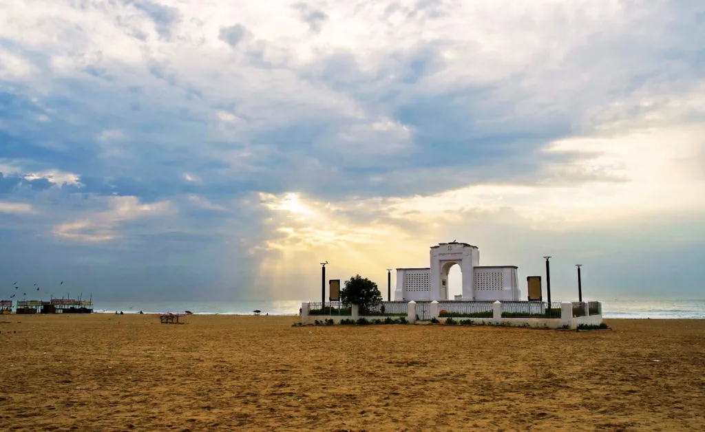 A serene beach scene at sunrise with soft sunlight peeking through clouds, casting a warm glow on a white monument. The mood is peaceful and contemplative.