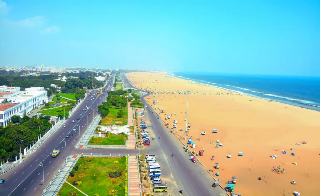 Aerial view of a vast sandy beach beside a bustling coastal road lined with buildings and green spaces. The blue ocean meets a clear sky, reflecting a serene, vibrant atmosphere.