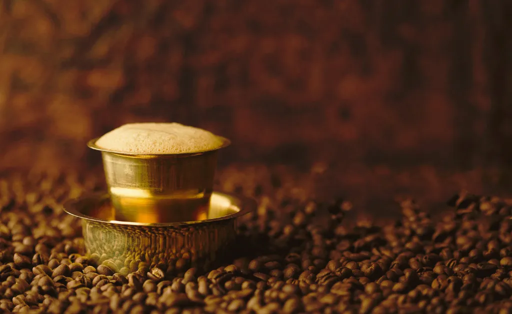 A frothy coffee in a traditional brass cup and saucer is surrounded by coffee beans. The warm lighting creates an inviting and aromatic atmosphere.