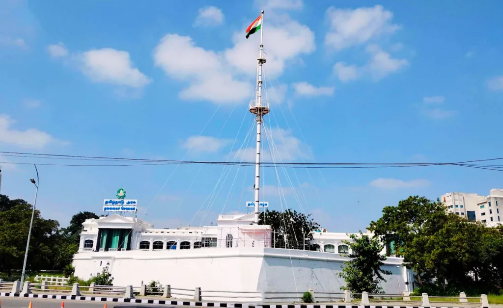 White building with an Indian flag atop a tall mast, set against a blue sky with fluffy clouds. Green trees surround the structure, creating a serene atmosphere.