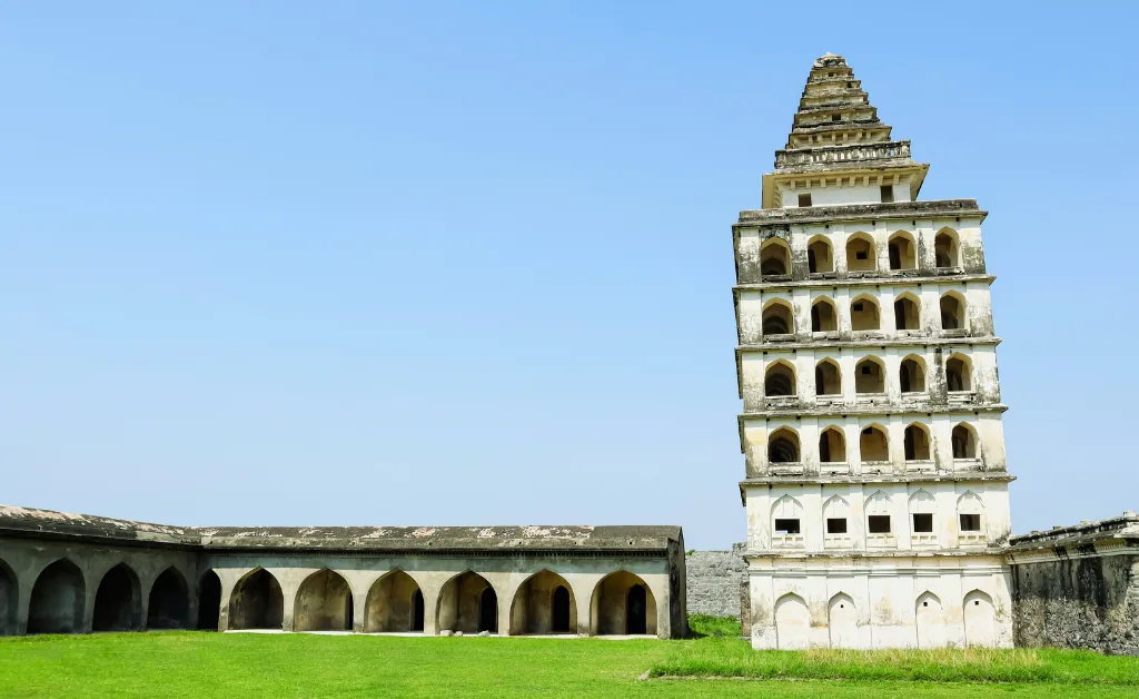 Ancient tiered tower with arches stands on lush green grass beside a long, arched building. The clear blue sky adds a serene backdrop.