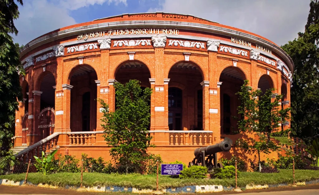 Red-brick, semi-circular building with archways, labeled "Museum Theatre." Surrounded by greenery, a historic cannon in front conveys heritage.