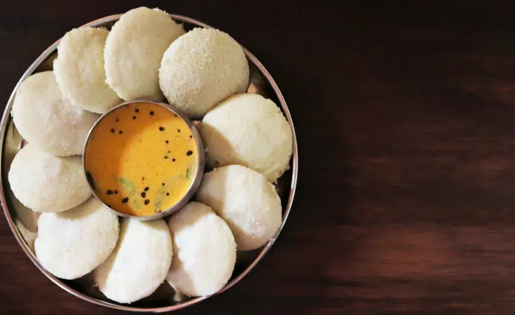 A plate of fluffy, round idlis arranged in a circle around a small bowl of orange coconut chutney, set on a dark wooden table, conveying a warm, inviting tone.