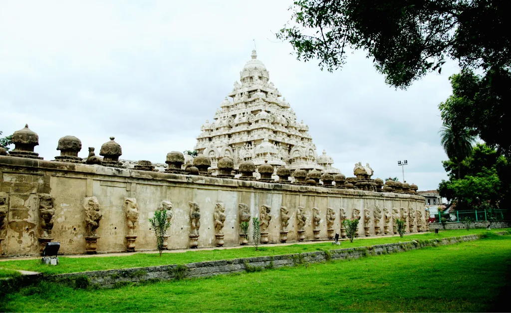 Ancient temple with intricate carvings stands tall behind a stone wall adorned with statues. Green grass and trees frame the scene under a cloudy sky.