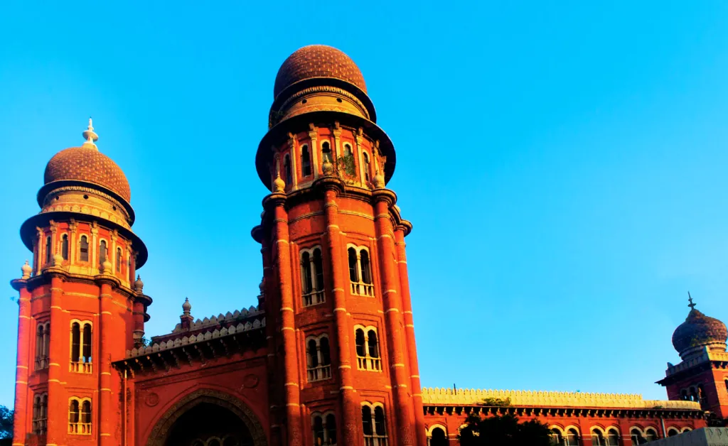 A historic red-brick building with two prominent domed towers against a clear blue sky. The building's ornate architecture evokes grandeur and elegance.