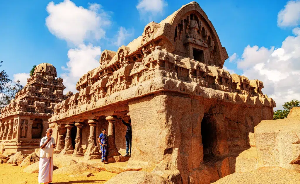 Ancient stone temple with intricate carvings and majestic arches under a clear blue sky. Tourists in traditional attire explore, evoking a sense of history.