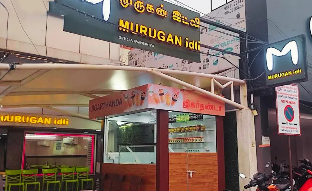 Brightly lit entrance of Murugan Idli Shop with vibrant signage in Tamil and English. Interior has green chairs. Scooters are parked outside.
