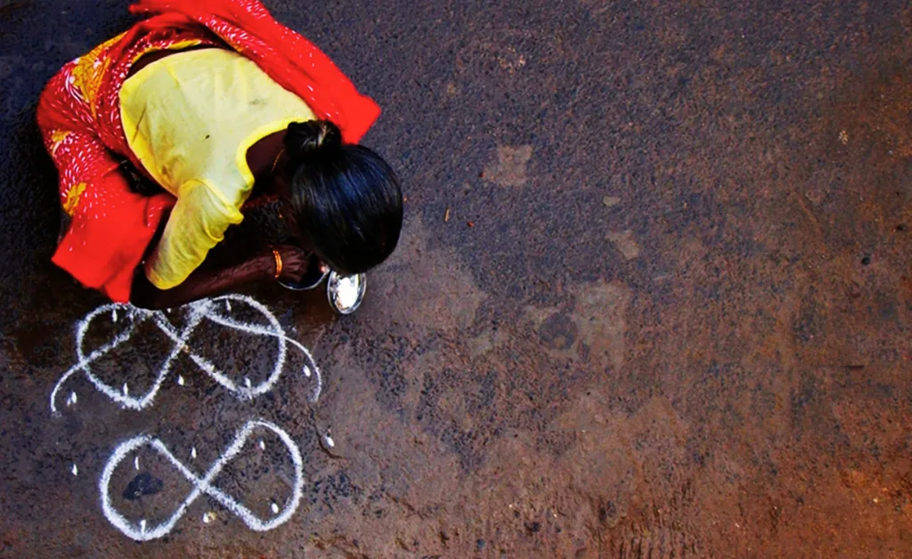 A woman in a red and yellow sari draws intricate white rangoli patterns on a dusty street, using powder and a bowl, conveying cultural artistry and tradition.