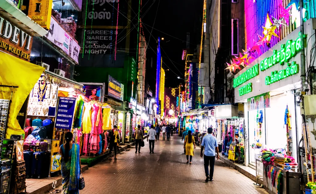 A vibrant, bustling street market at night, illuminated by colorful neon lights. Shops display bright textiles and garments. People walk, creating a lively, energetic atmosphere.