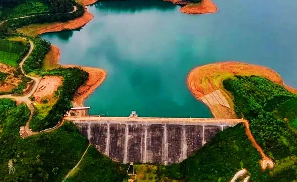 An aerial view of a large dam holding back a turquoise reservoir, surrounded by lush green hills and patches of brown earth, conveying a serene landscape.