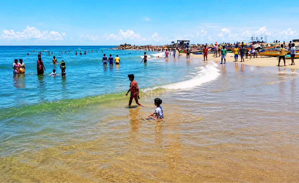 A crowded beach scene under a bright blue sky, with families and children playing in the shallow waves. Colorful clothes and boats on the sandy shore convey a lively atmosphere.