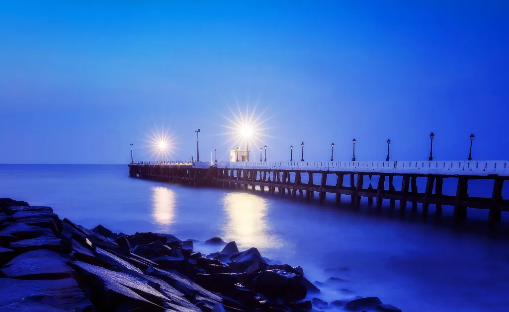 A serene pier at night with two bright lights reflecting on calm ocean water. Rocks line the shore, and the sky is a rich blue, creating a peaceful mood.