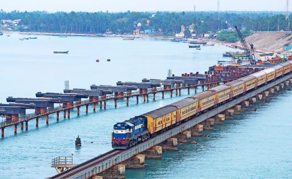 A train travels across a long bridge over calm blue water, with a distant shoreline lined with trees and buildings. The scene conveys tranquility.