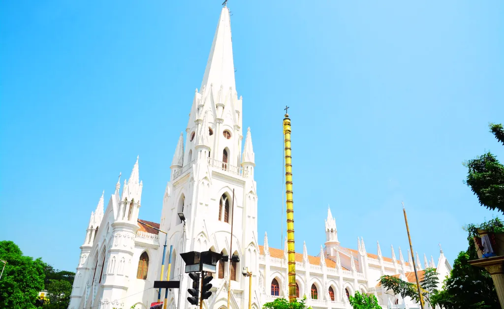 White Gothic-style cathedral with tall spire against a clear blue sky. Lush greenery frames the scene, conveying a serene and majestic atmosphere.