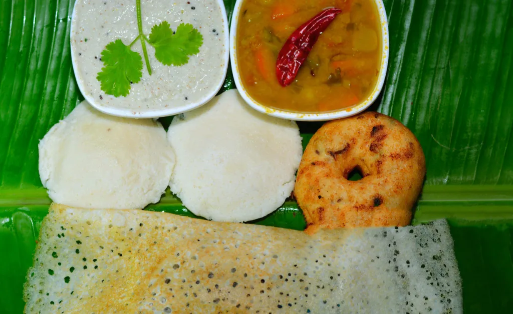A vibrant South Indian meal on a banana leaf features dosa, idli, vada, coconut chutney with coriander garnish, and sambar with a red chili, evoking a warm, traditional feel.