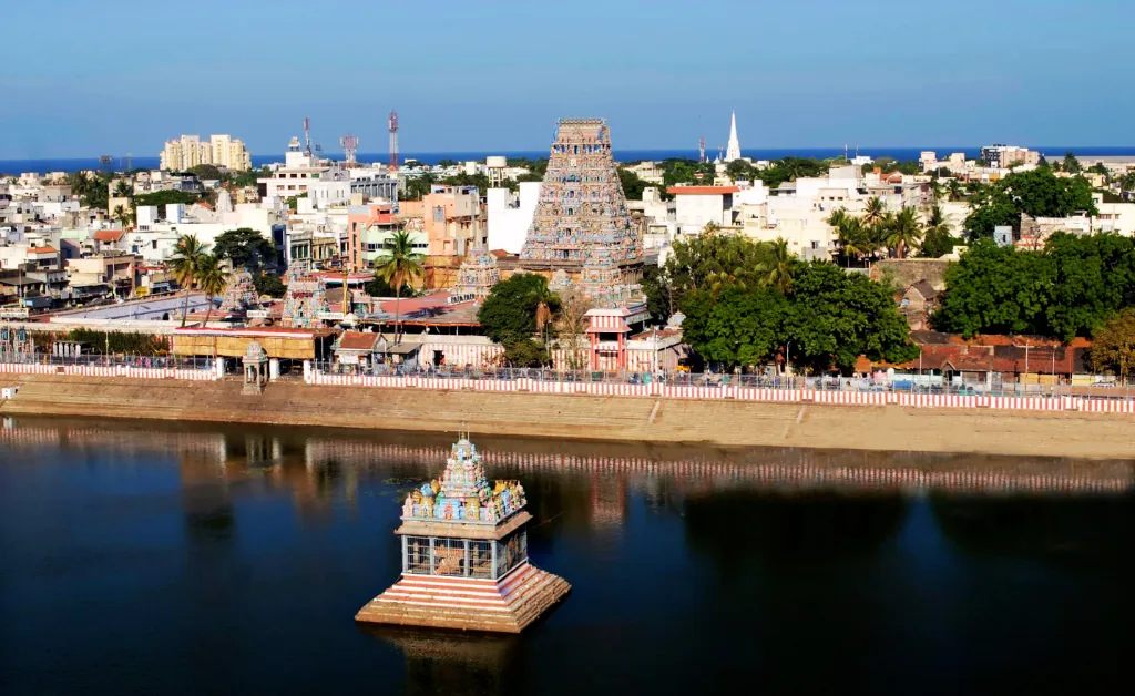 A serene cityscape featuring a colorful Hindu temple with intricate architecture, reflected in a tranquil water tank, surrounded by lush greenery and urban buildings under a clear blue sky.
