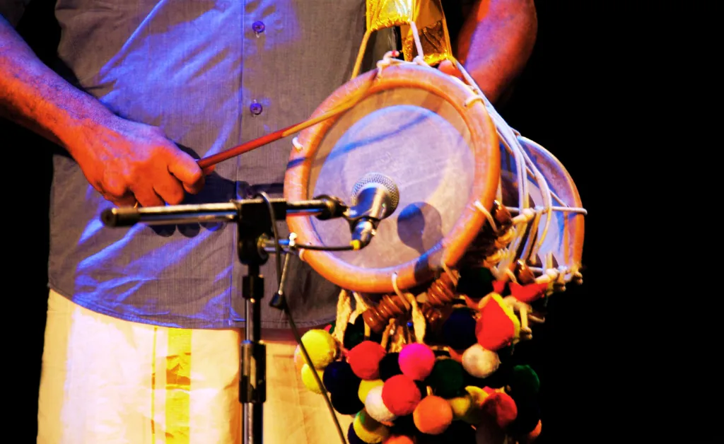 A person plays a traditional drum, possibly a damaru, in warm stage lighting. The drum is adorned with colorful pom-poms, creating a lively atmosphere.