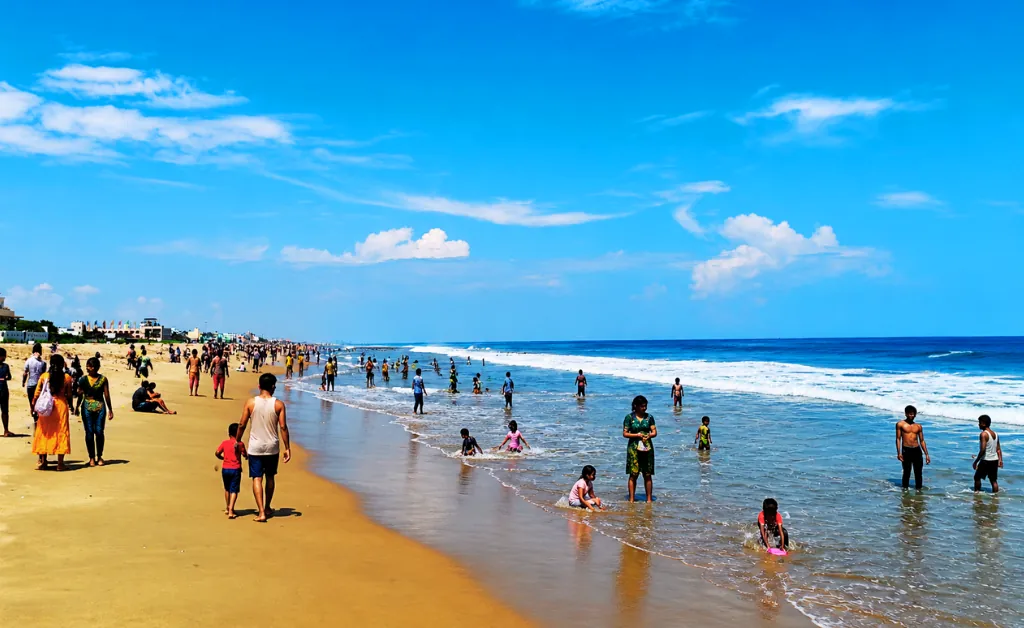 Crowded beach scene under clear blue skies. People of all ages enjoy walking, swimming, and playing near the shoreline. Energetic and sunny atmosphere.