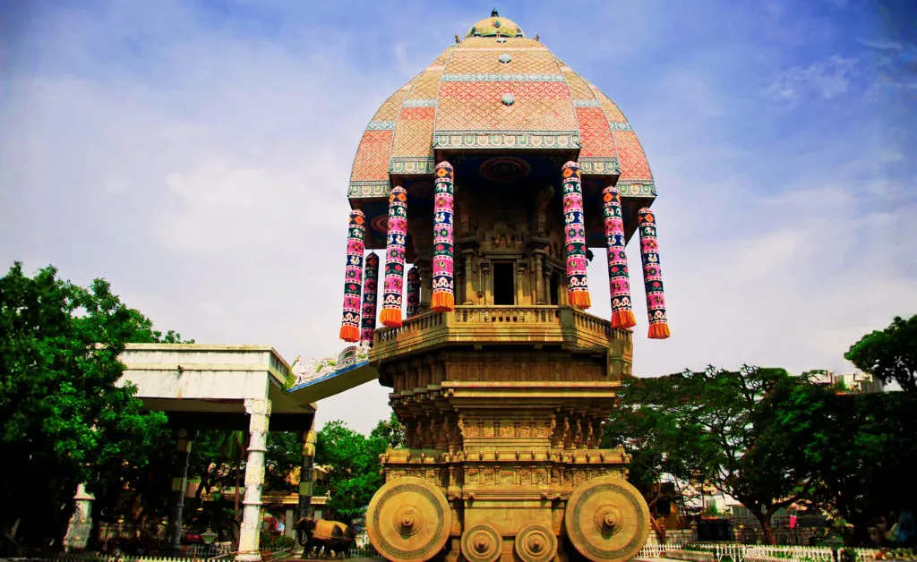A majestic, intricately decorated temple chariot with a domed roof and vibrant pillars stands prominently, surrounded by greenery and a vibrant blue sky.