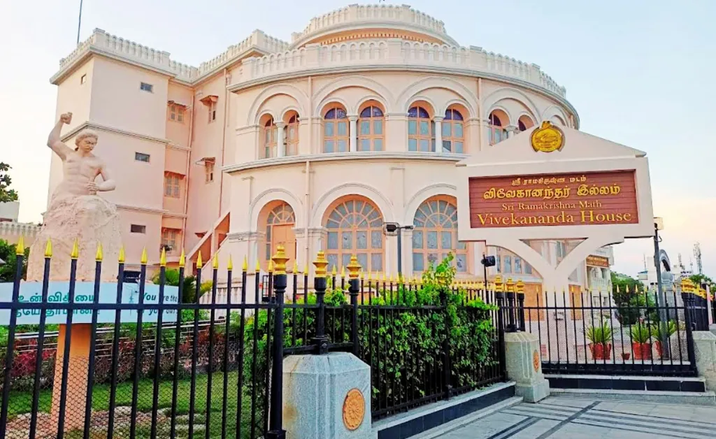 A historic building with arched windows and ornate details, known as Vivekananda House, features a Swami Vivekananda statue outside, surrounded by a black iron fence. The atmosphere is serene and culturally rich.